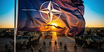NATO flag waving with military exercise in background, symbolizing alliance strength.
